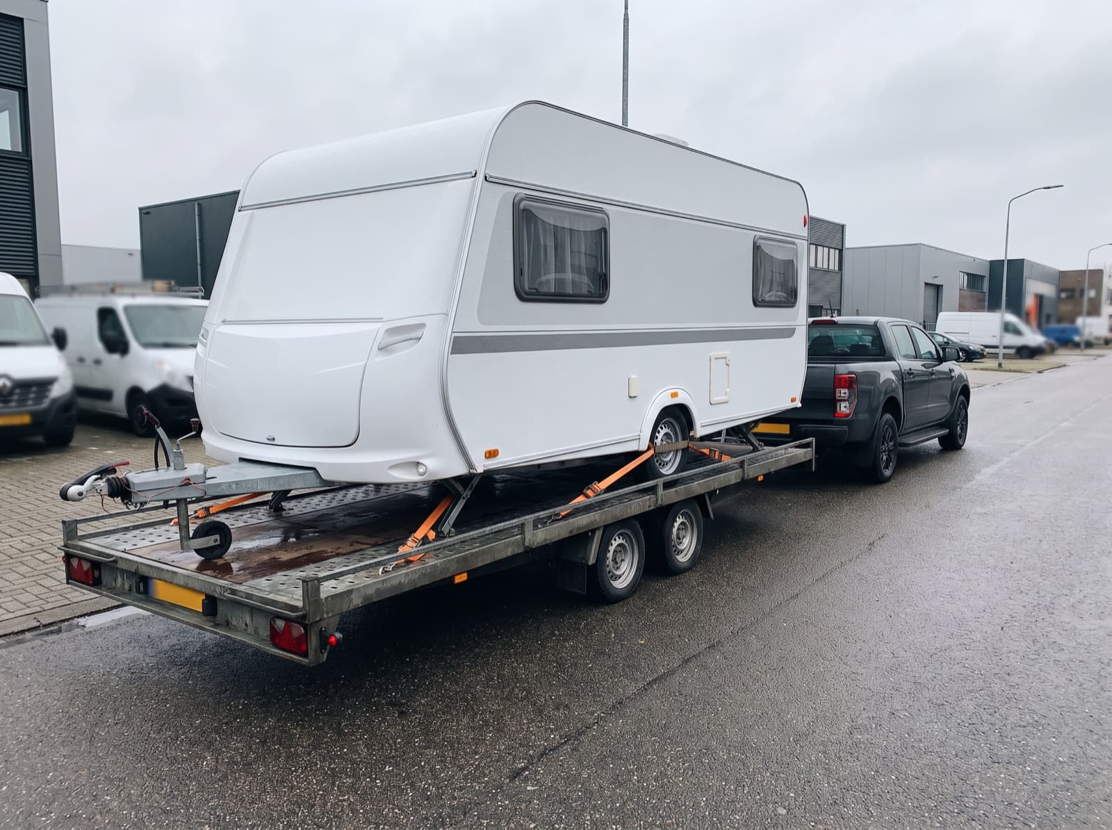 Witte caravan vastgezet op een open trailer achter een pick-up op een bedrijventerrein